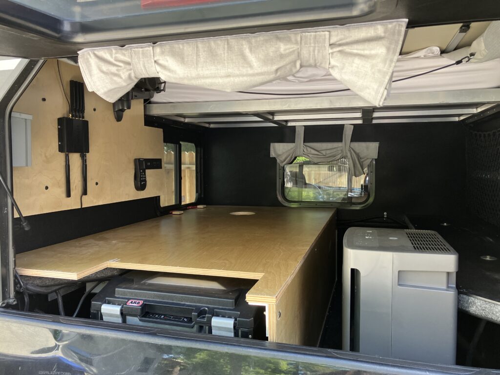Wide interior view of the Project M build showing the third bed plywood platform, ARB fridge in its slide below, and the GE dehumidifier